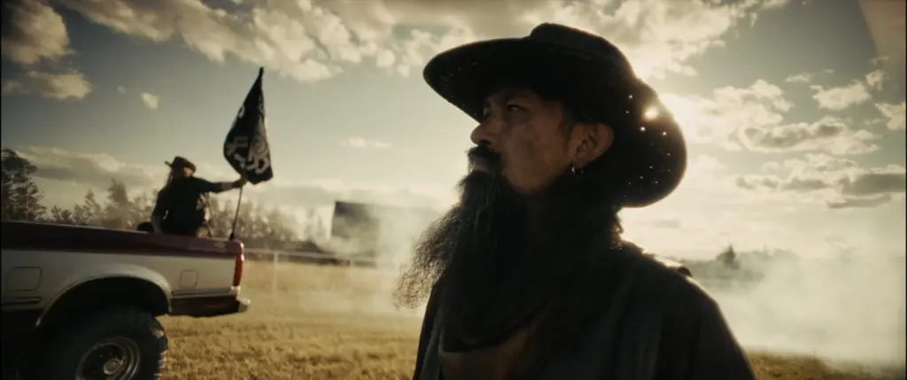Profile of a long-bearded man in a cowboy hat; behind him a rider waves a black flag from a pickup at golden hour - Amon Amarth