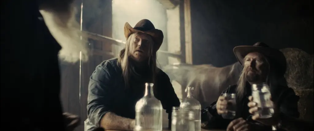 Two long-haired men in cowboy hats at a table with glass bottles inside a dusty, backlit barn - Amon Amarth
