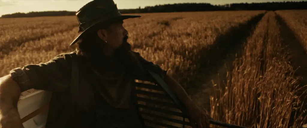 Bearded man in a cowboy hat riding on a moving vehicle beside a golden wheat field at sunset - Amon Amarth