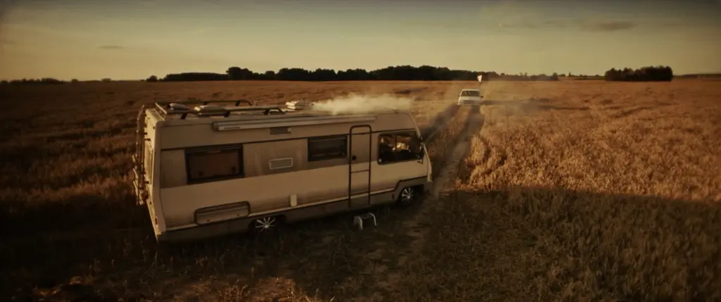 A vintage motorhome parked at the edge of a wheat field at golden hour, a car driving off in the distance - Amon Amarth