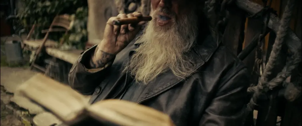 Close-up of a bearded old man in a worn leather coat drawing on a cigar, tattooed hand raised - Amon Amarth
