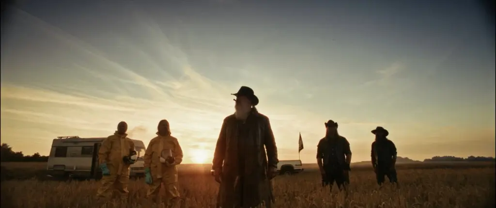 Silhouetted group - two in hazmat suits, a long-coated leader and cowboys - before a low sun in a field - Amon Amarth