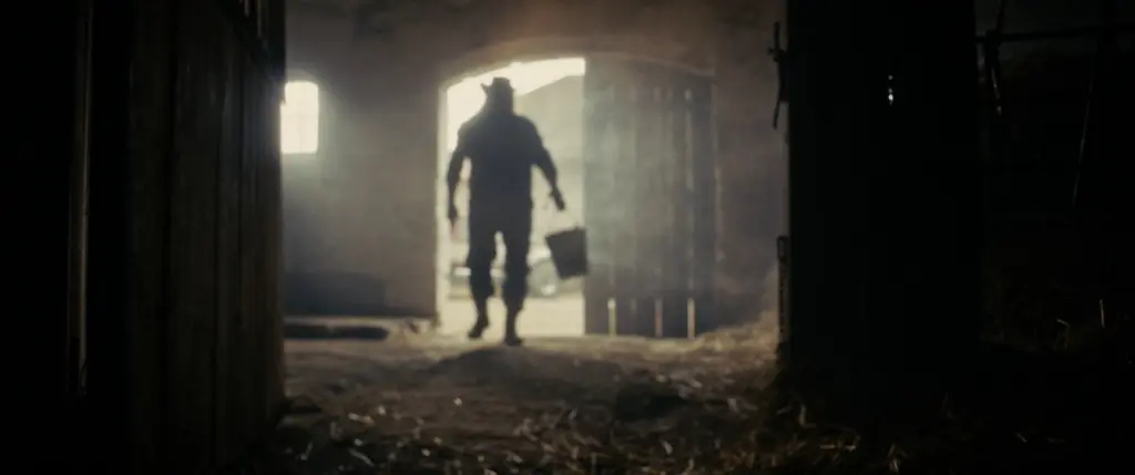 Silhouette of a figure in a cowboy hat carrying a bucket through a smoky, backlit barn doorway - Amon Amarth