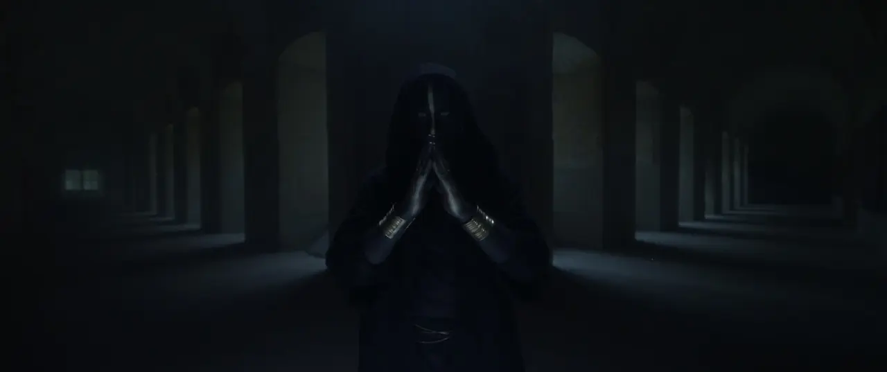 IGORRR music video still - hooded figure with painted face, hands pressed together in a candlelit colonnade