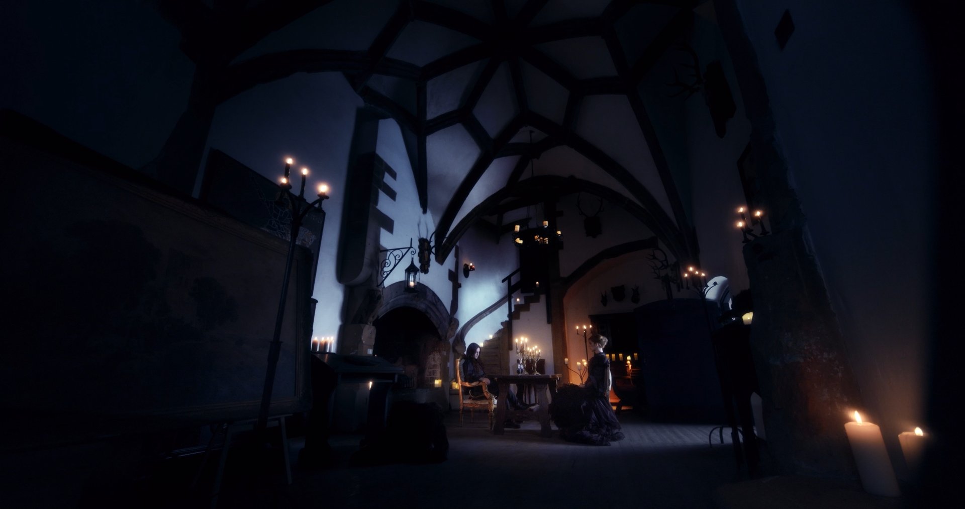 Ater music video still - wide shot of a vaulted stone hall, two figures seated at a candlelit table