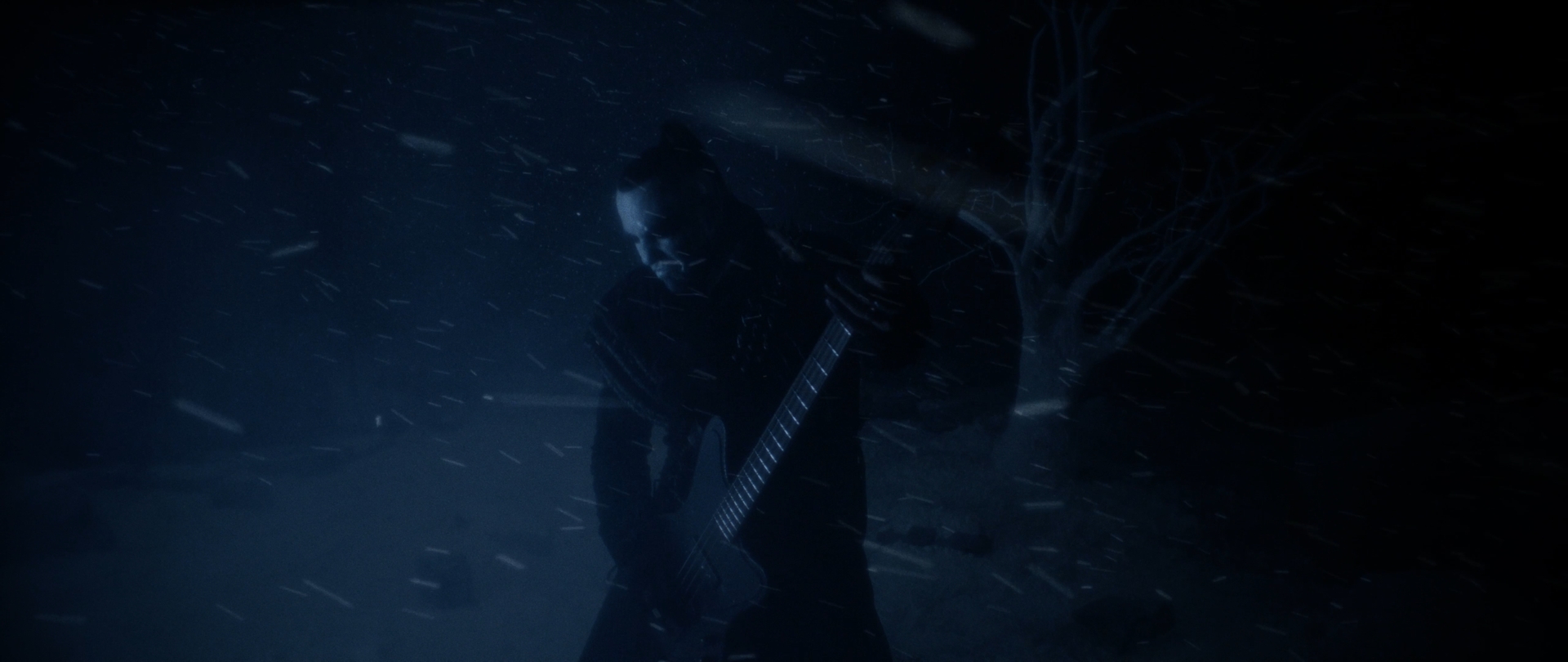 Behemoth Mystic still - bearded musician silhouetted with a guitar in a blizzard beside a bare tree
