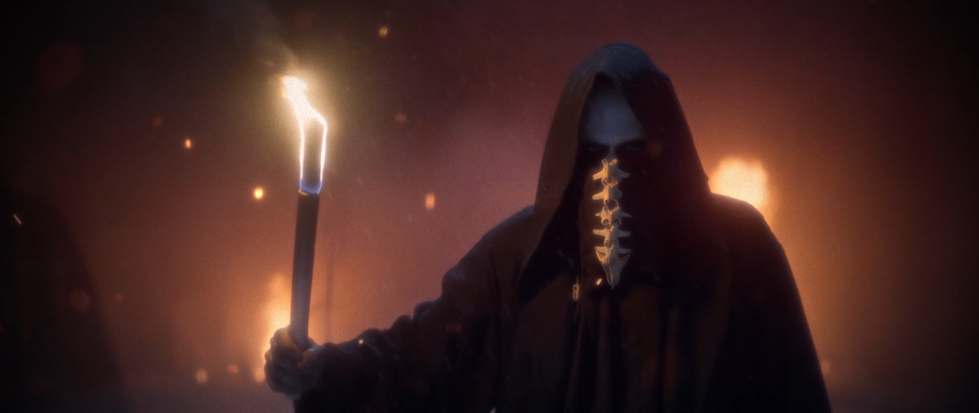 Behemoth Mystic still - hooded figure in a bone mask holding a lit torch, smoke and firelight behind