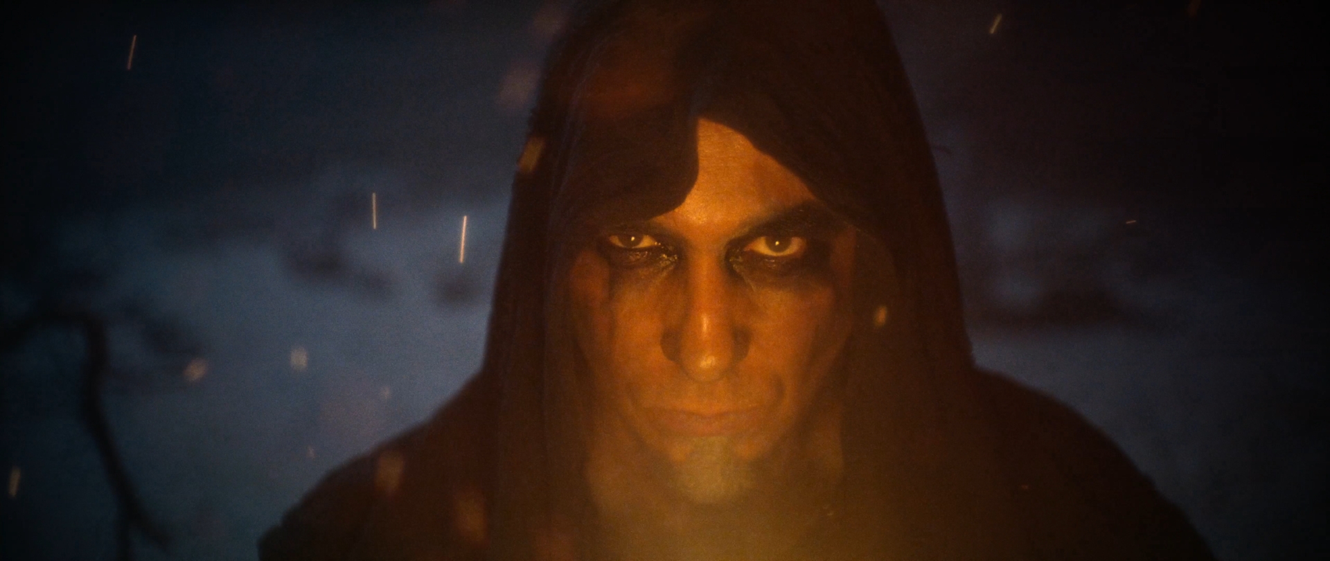 Behemoth Mystic still - extreme close-up of a hooded figure's face lit by firelight, embers drifting past