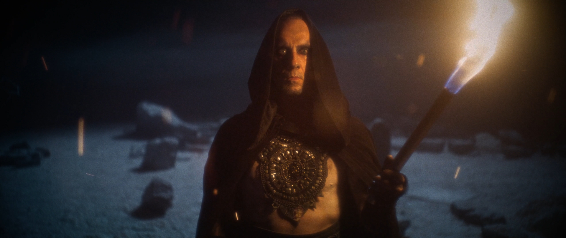 Behemoth Mystic still - hooded figure in a jeweled breastplate holding a lit torch among snow-covered standing stones
