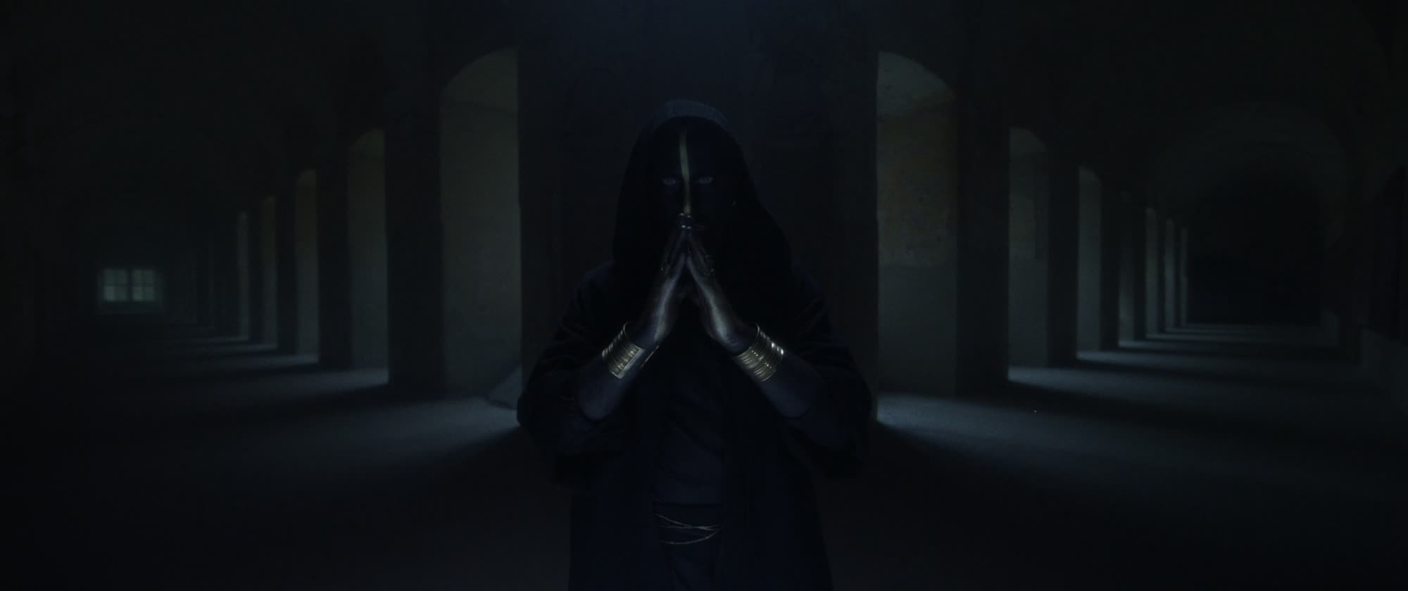 IGORRR music video still - hooded figure with painted face, hands pressed together in a candlelit colonnade