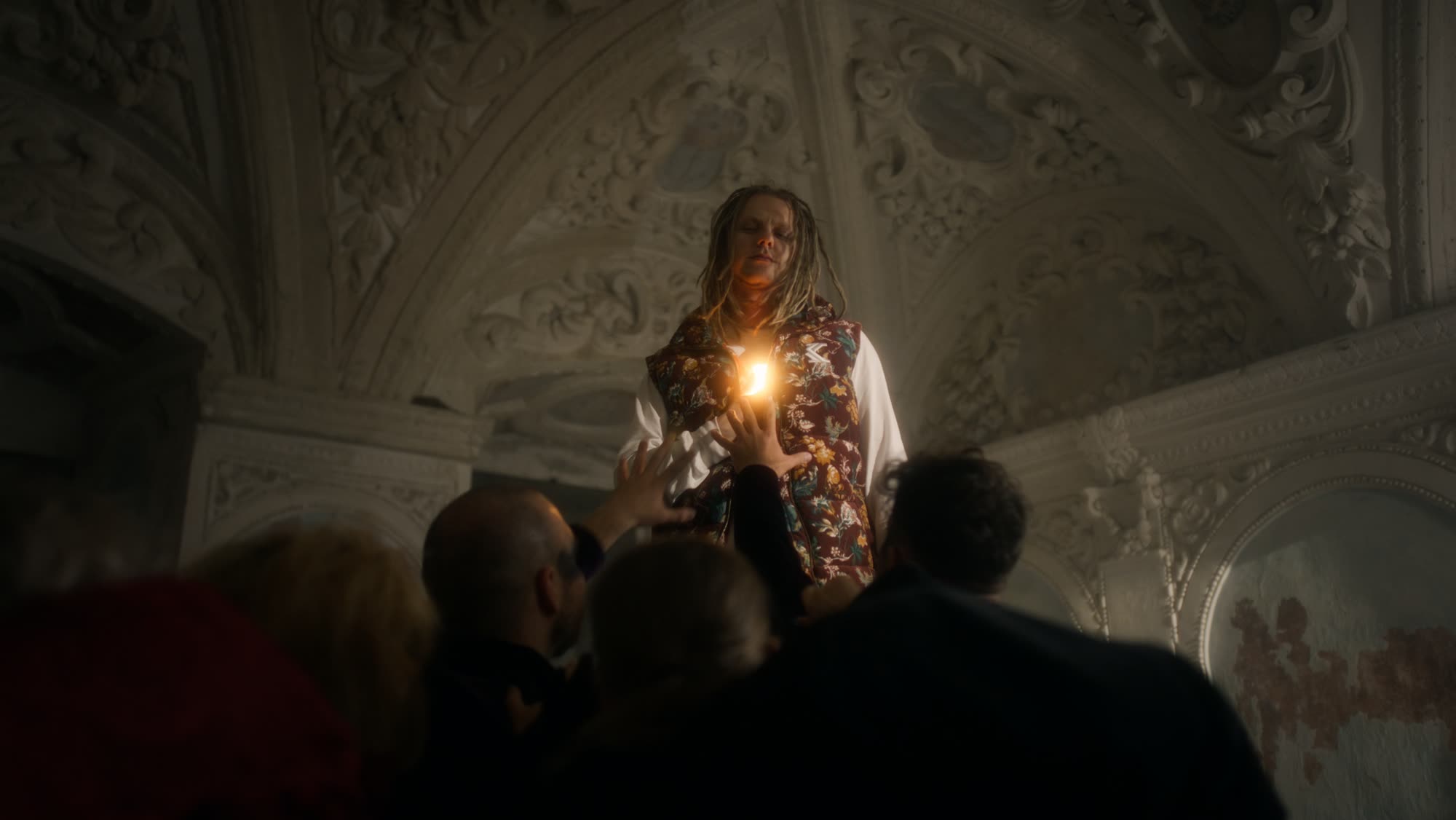 Jestem Alpha still - man in a floral folk vest holding a lit candle above a crowd reaching up toward him