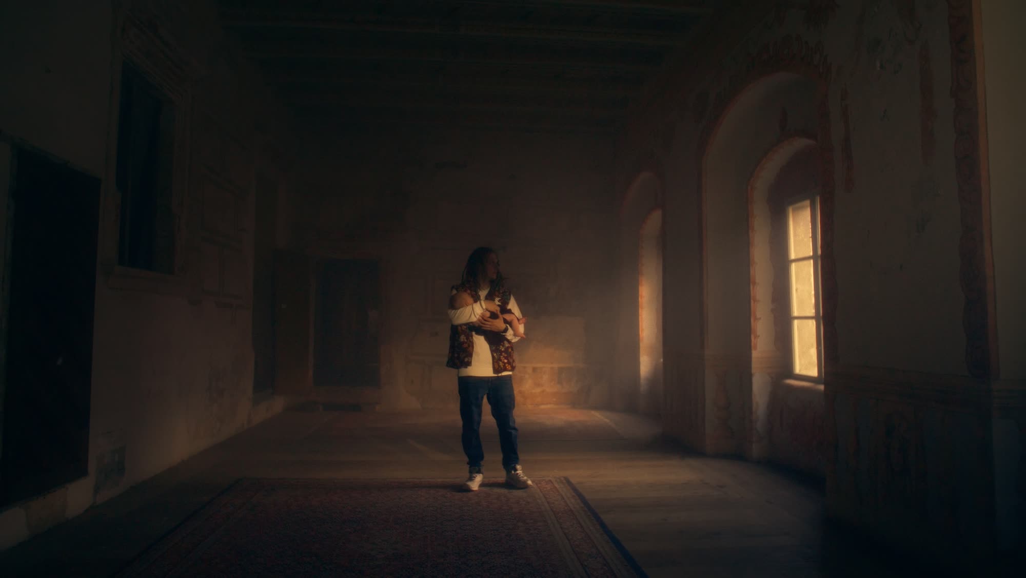 Jestem Alpha still - man carrying a baby, walking through a vast decaying palace hall