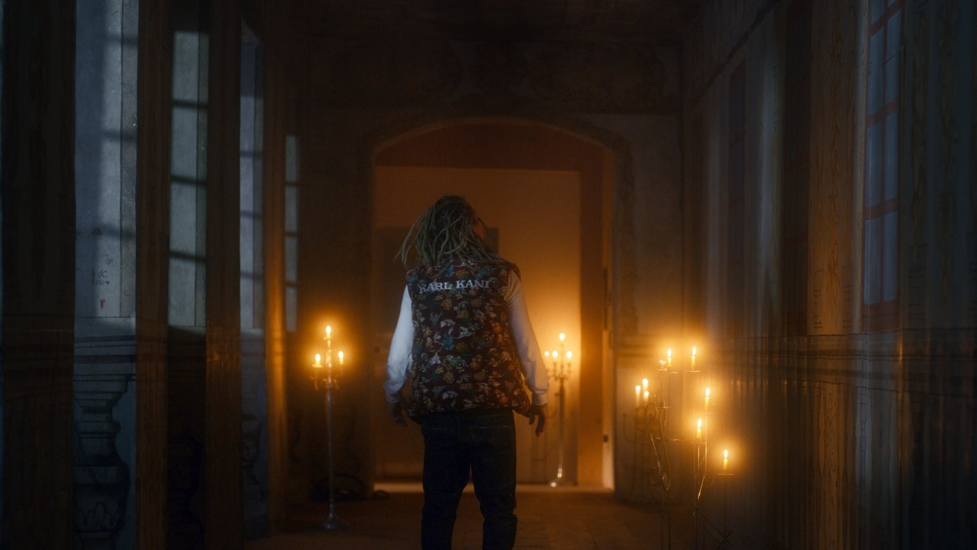 Jestem Alpha still - figure in a floral folk vest walking down a candlelit corridor