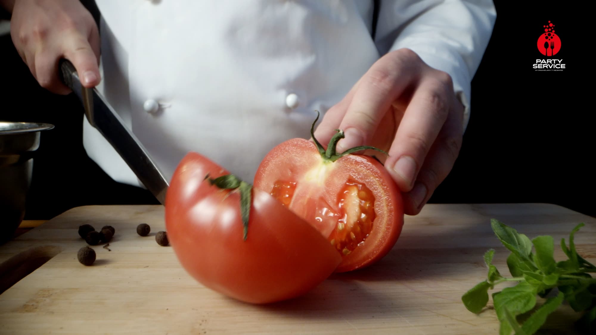 Chef slicing a tomato in half on a wooden board, herbs and peppers scattered around - Party Service commercial
