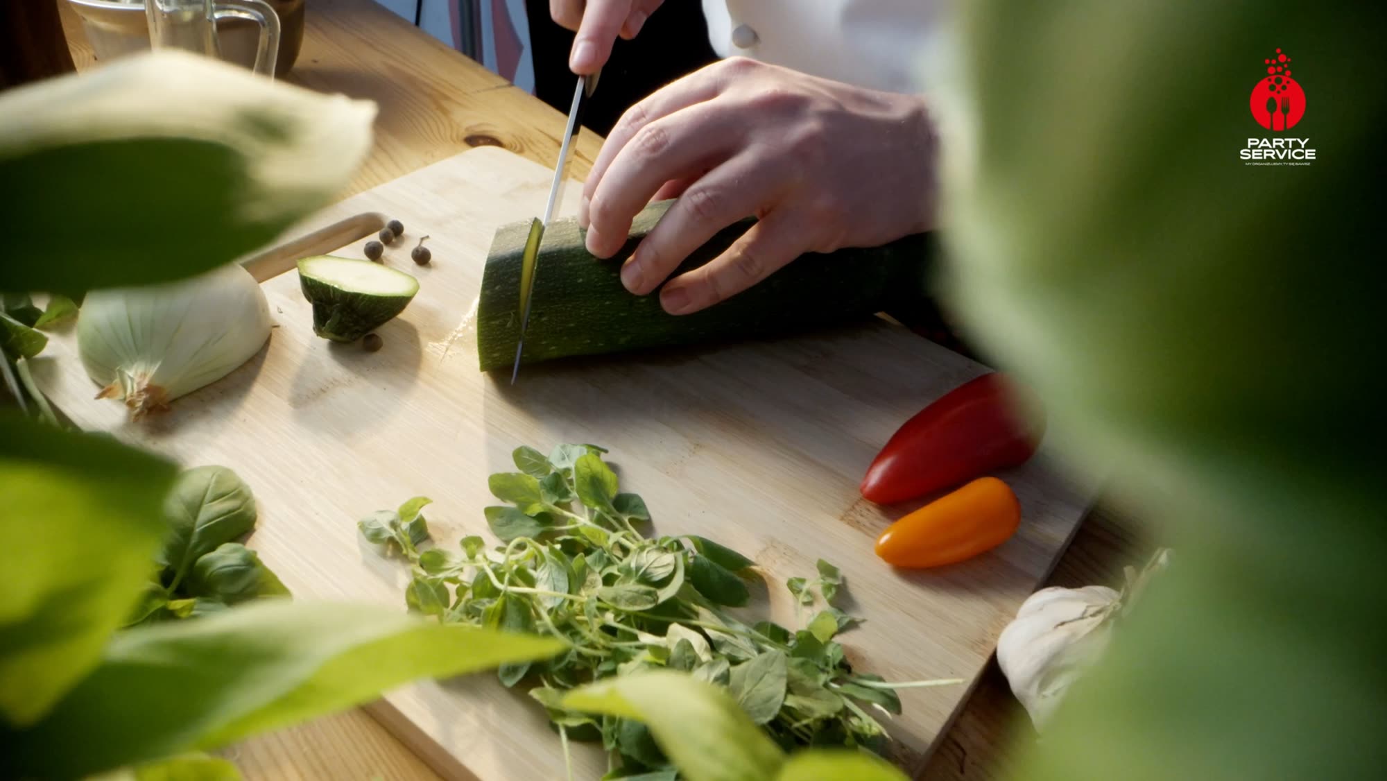 Chef trimming courgette on a wooden board surrounded by fresh herbs and vegetables - Party Service commercial