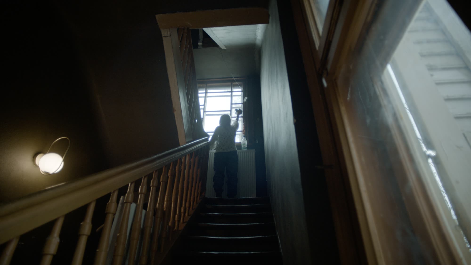 Low silhouette on a staircase, hand raised toward a bright window at the top