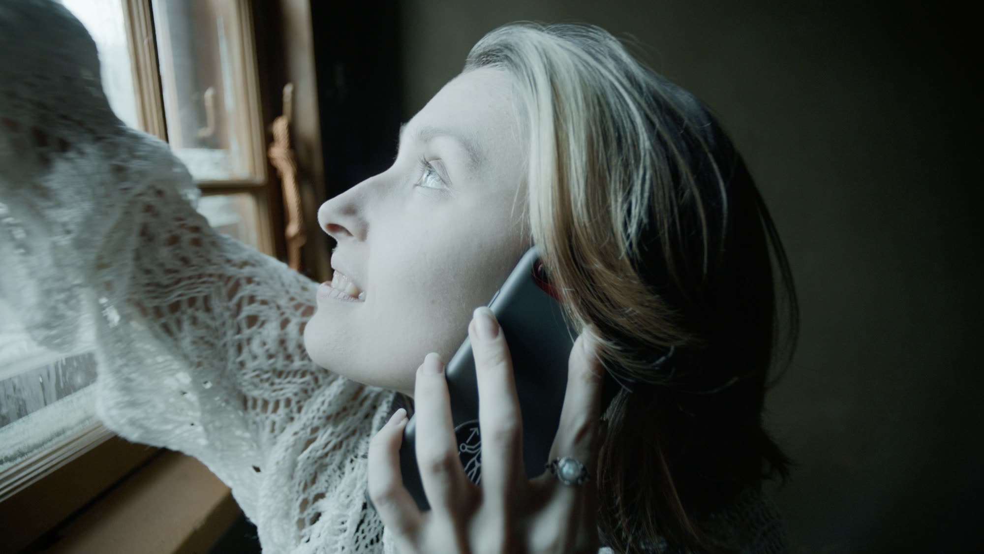 Woman reaching toward a frosted window while on a phone call, lit only by winter daylight