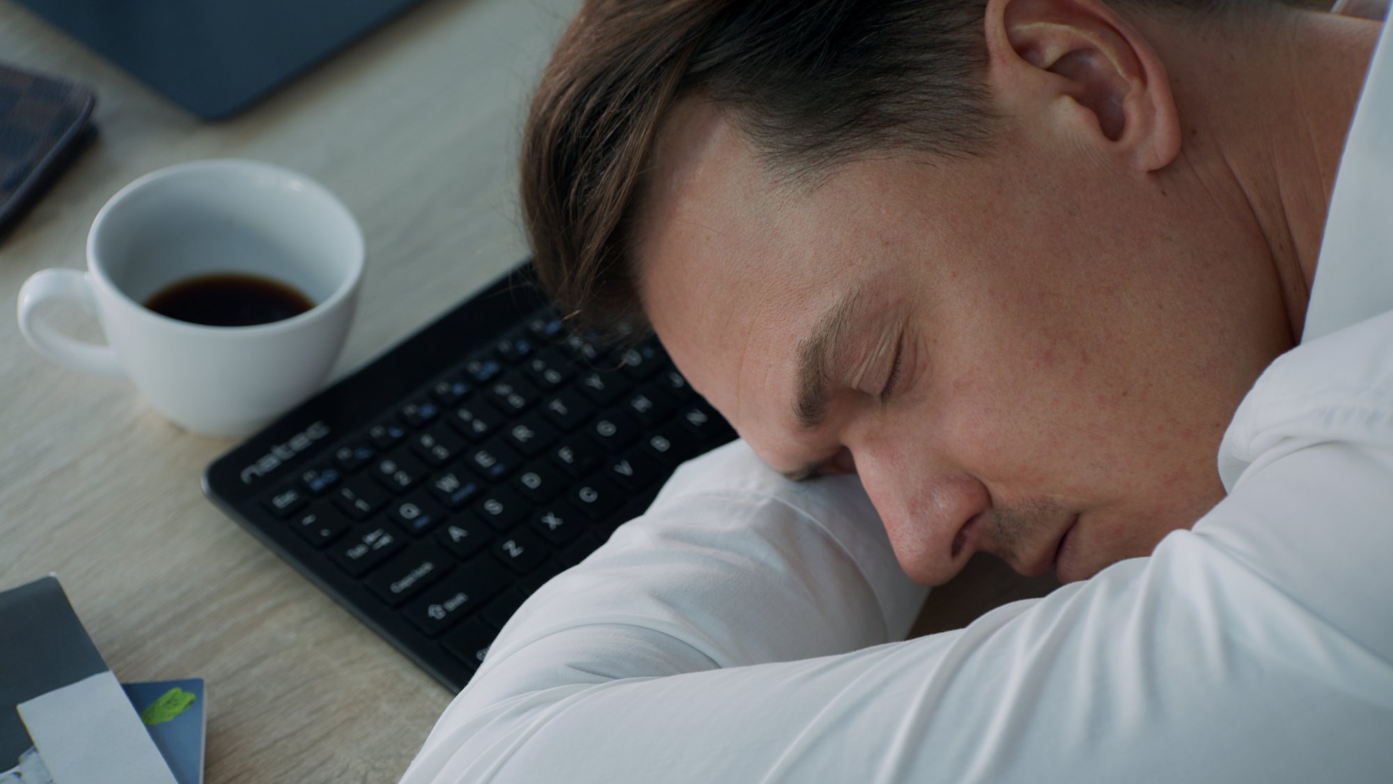 Man asleep at his office desk, head down beside the keyboard and a coffee cup - Plantpur commercial