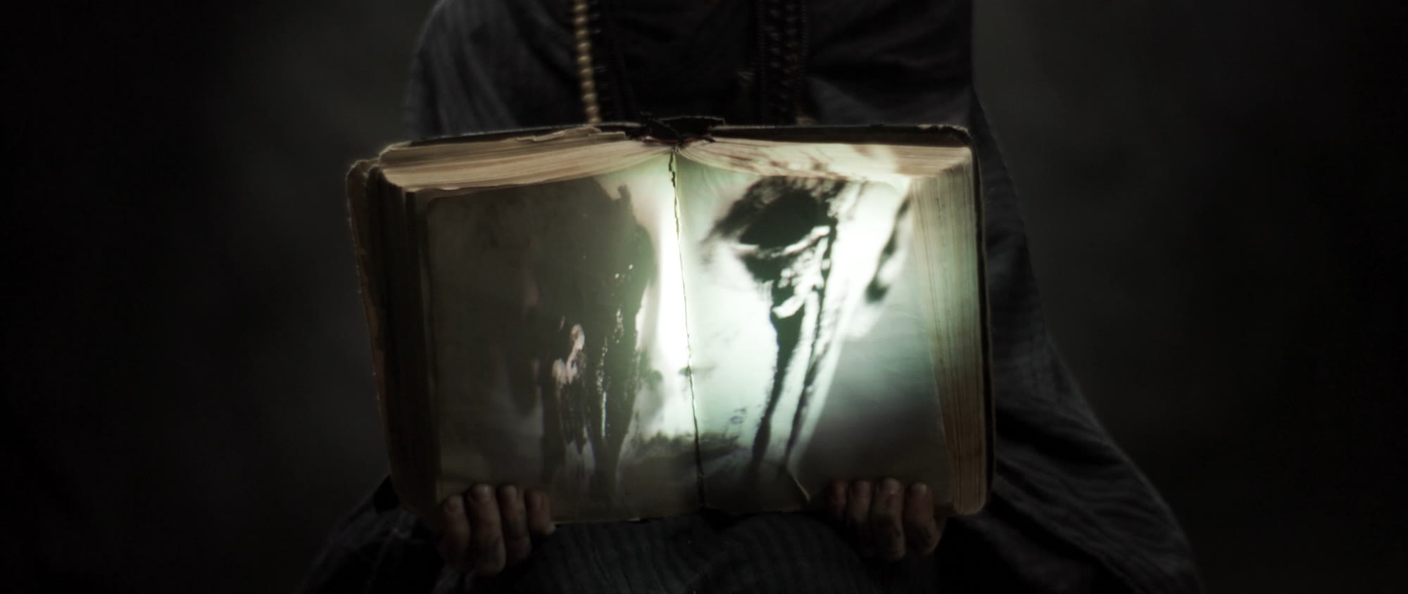 Behemoth music video still - hooded figure holding an open book with a ghostly photograph inside