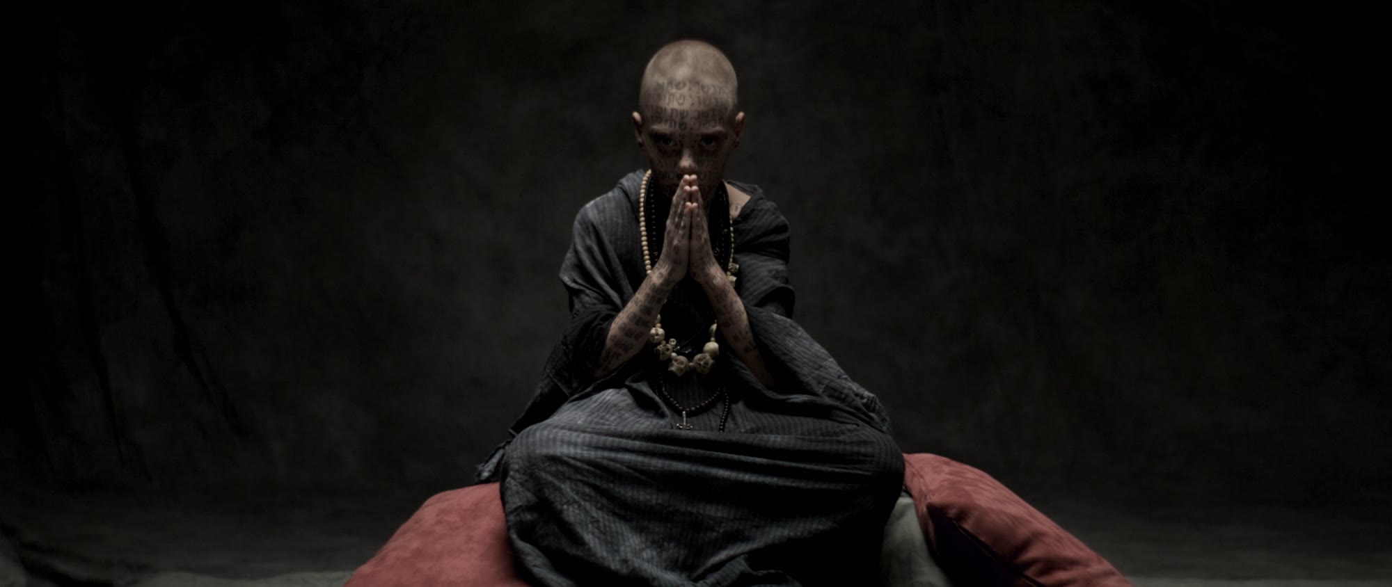 Behemoth music video still - tattooed bald figure seated, hands pressed together in prayer
