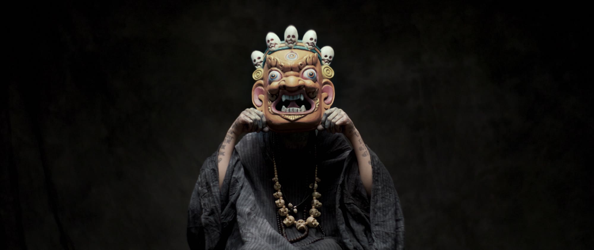 Behemoth music video still - figure holding a painted ceremonial mask crowned with small skulls