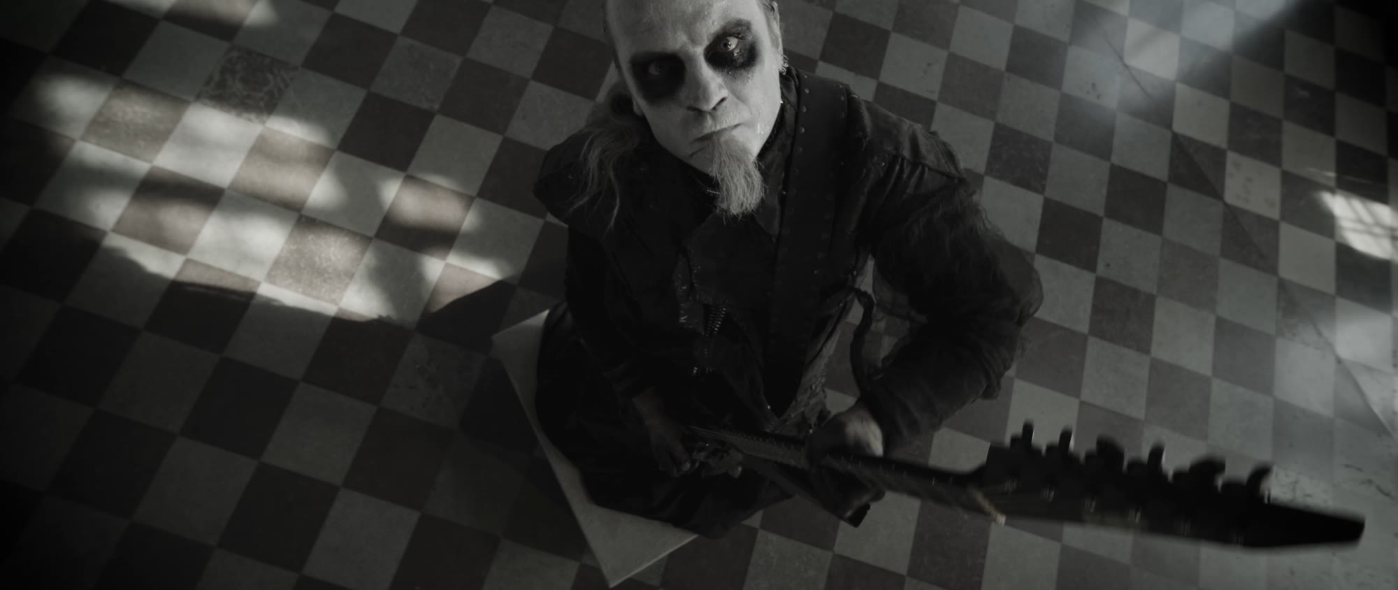 Behemoth music video still - bearded musician seated on a checkered floor holding a guitar