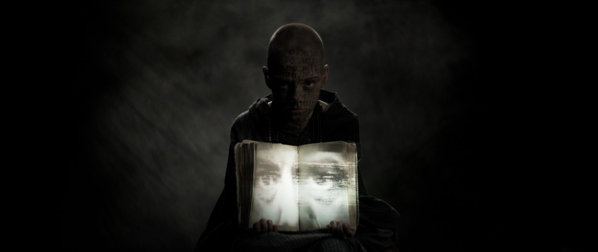 Behemoth music video still - hooded figure standing before a glowing screen showing a ghostly face