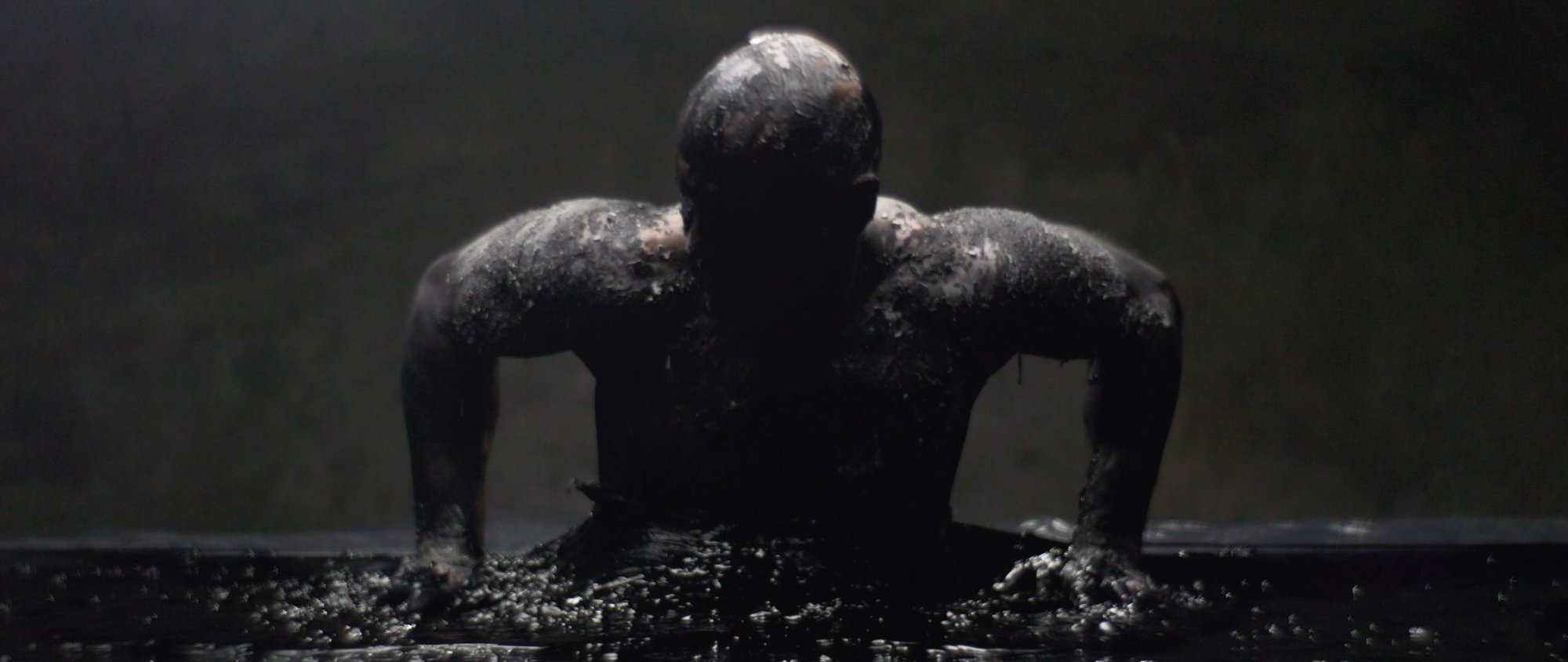 Behemoth music video still - muscular figure reaching a hand up from a bed of dark clay