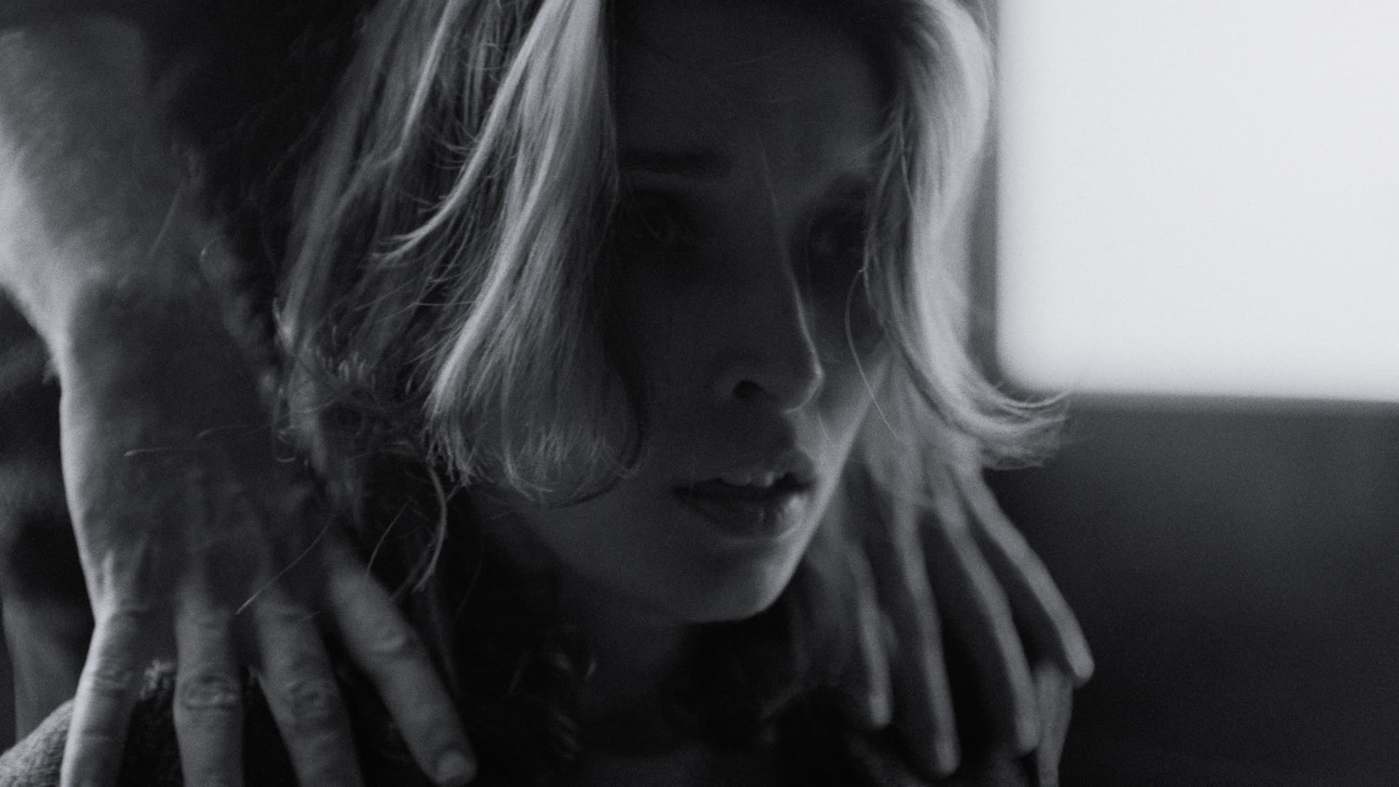 Close-up on her face, a blurred figure standing in the distance behind glass