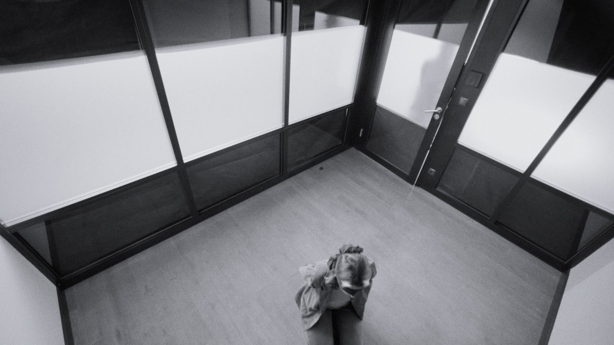 Overhead shot of a figure curled small on the floor of an empty glass-walled office corridor