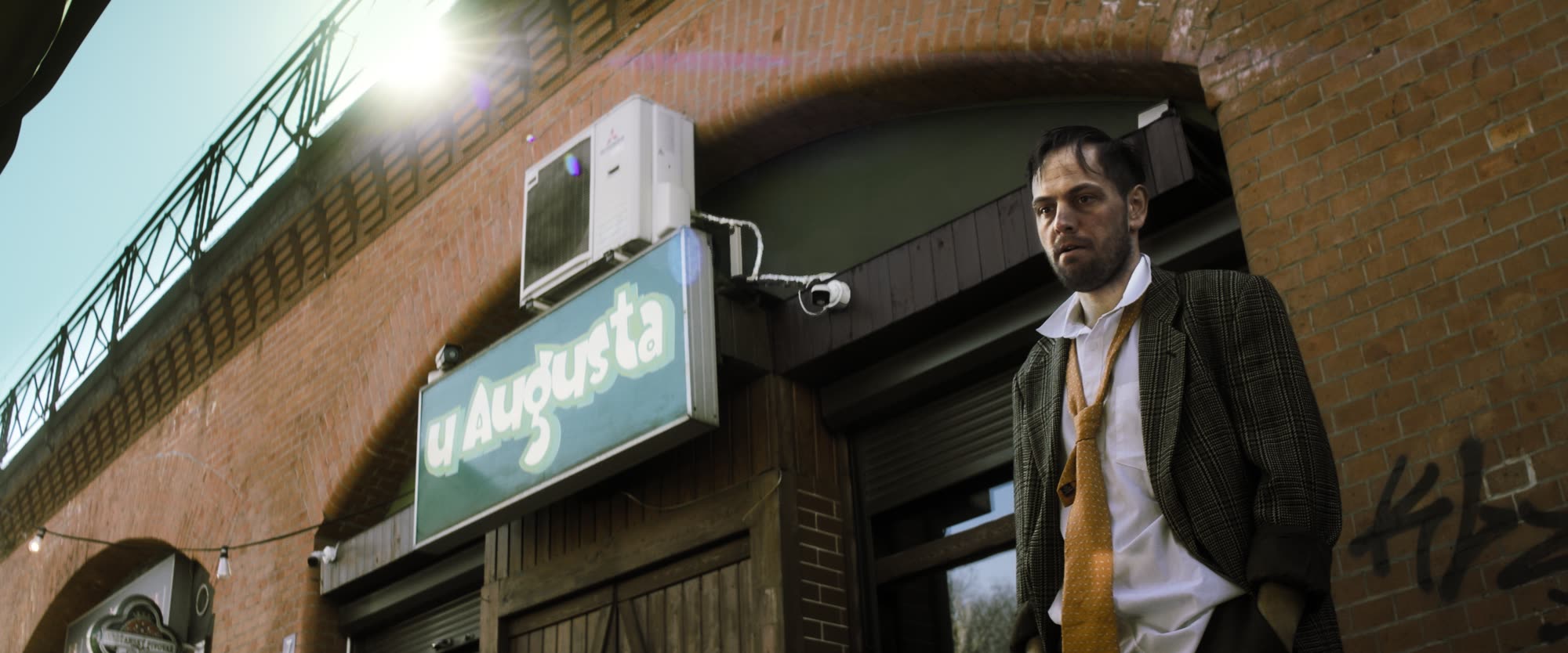 Man in a suit and loosened tie standing beneath a bar sign, backlit by low sun