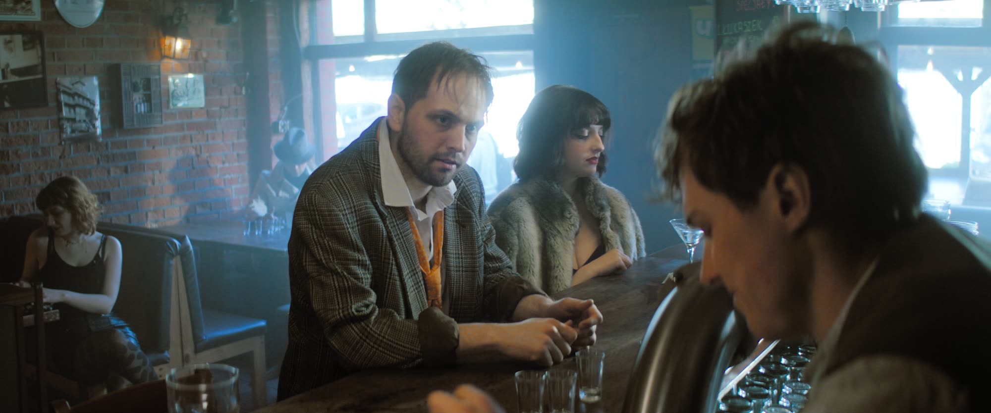 Man in a plaid jacket leaning on a bar counter beside a woman in a fur stole, smoky retro-lit bar